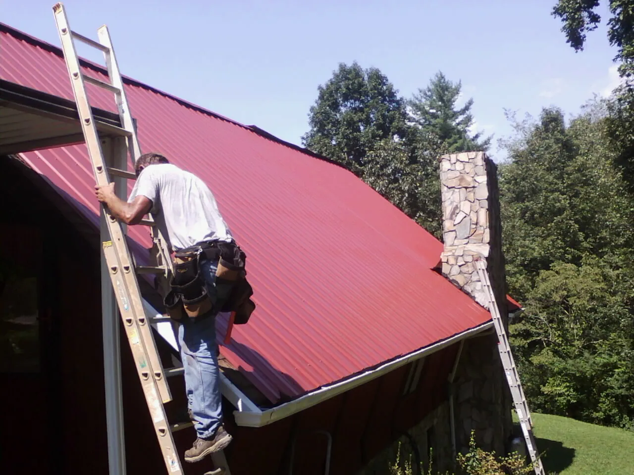 Roofer on a ladder beside a red metal roof and stone chimney — Soffit & Fascia Repair by Roofing Brothers in Gaylordsville